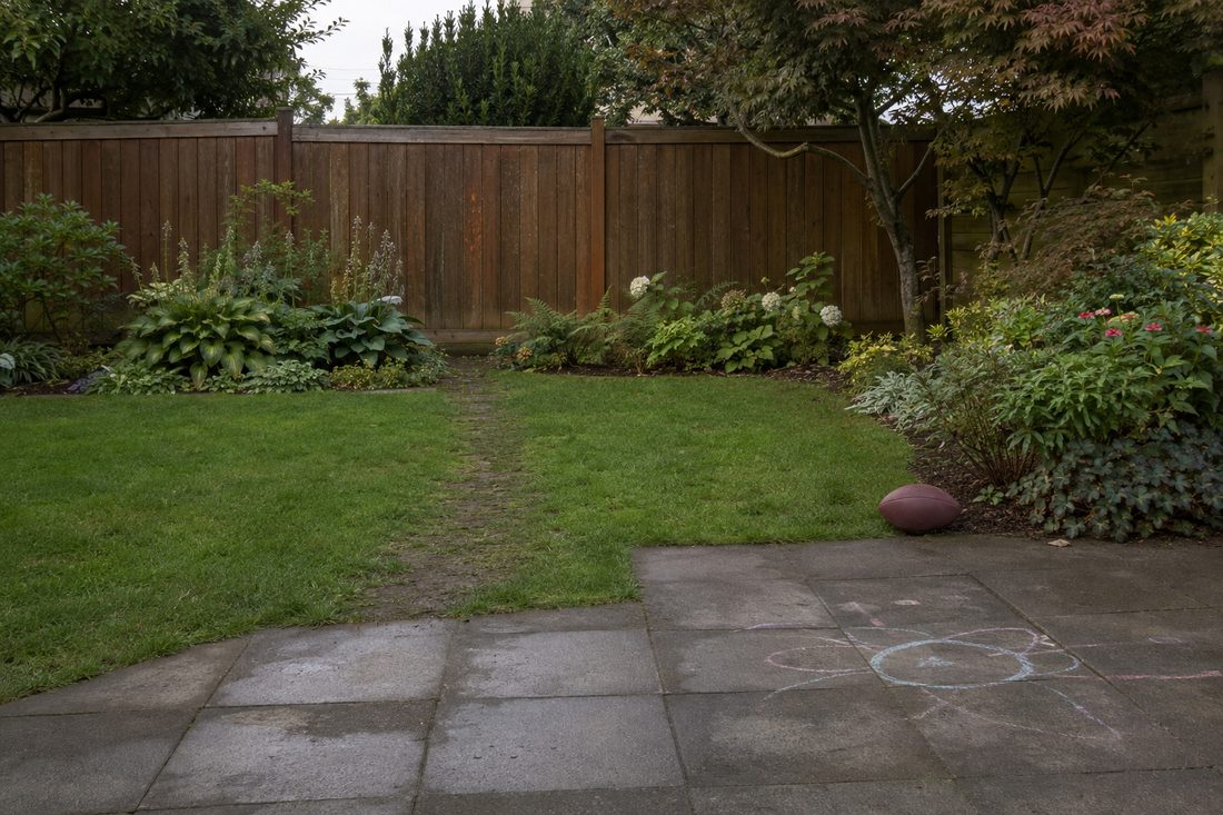 A small back lawn on an overcast afternoon with a football left near a flower bed and a faded chalk drawing on the patio stones.